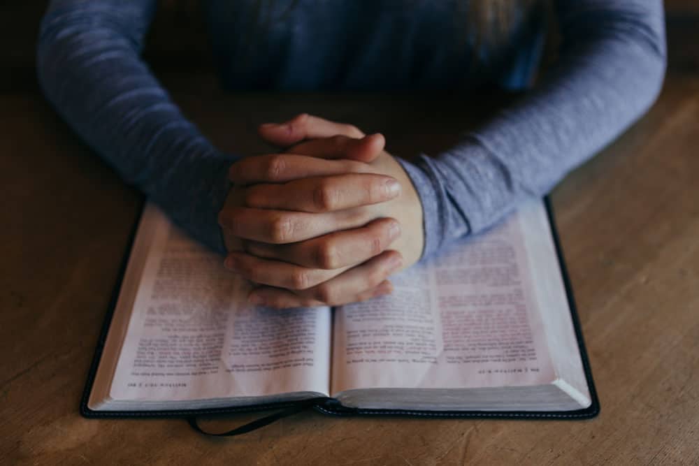 hands folded in prayer laying over an open Bible