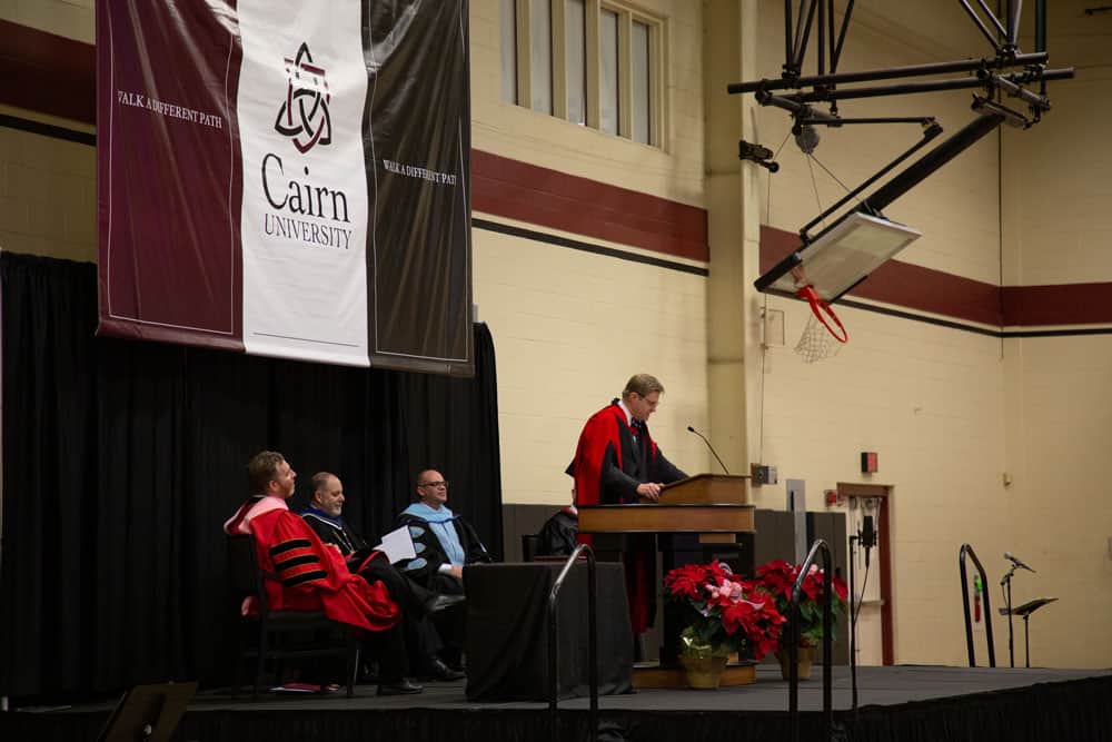 shot taking from side profile; Jonathan Master standing at podium on stage in academic regalia 