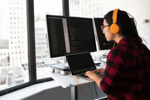 woman wearing headphones writing computer code on a computer facing window