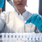 woman, sitting in front of a table with test tubes, is pictured from nose down adding a substance into a small test tube