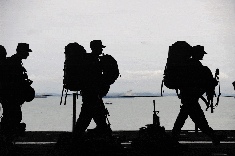 three soldiers in shadow walk with large backpacks across the image with a gray sky and sea behind them