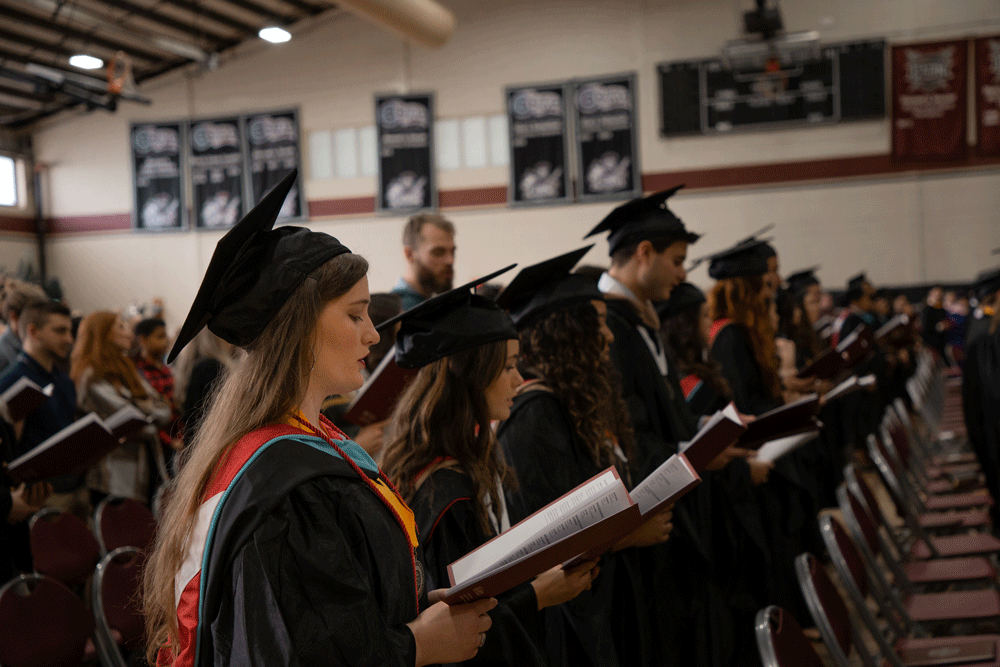 Side profile image of a row of graduates in their regalia, singing a hymn from the Commencement Program