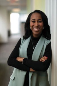 Breana Farrell wearing a gray vest and crossing her arms, leaning against an office wall, and smiling at the camera