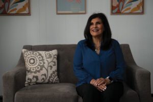 Paz Schroyer wearing a blue button-down shirt sitting on a gray couch, smiling and looking slightly off camera