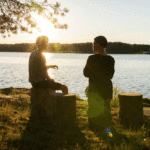 two people sitting next to a lake at sunset and talking