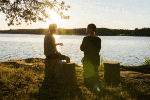 two people sitting next to a lake at sunset and talking