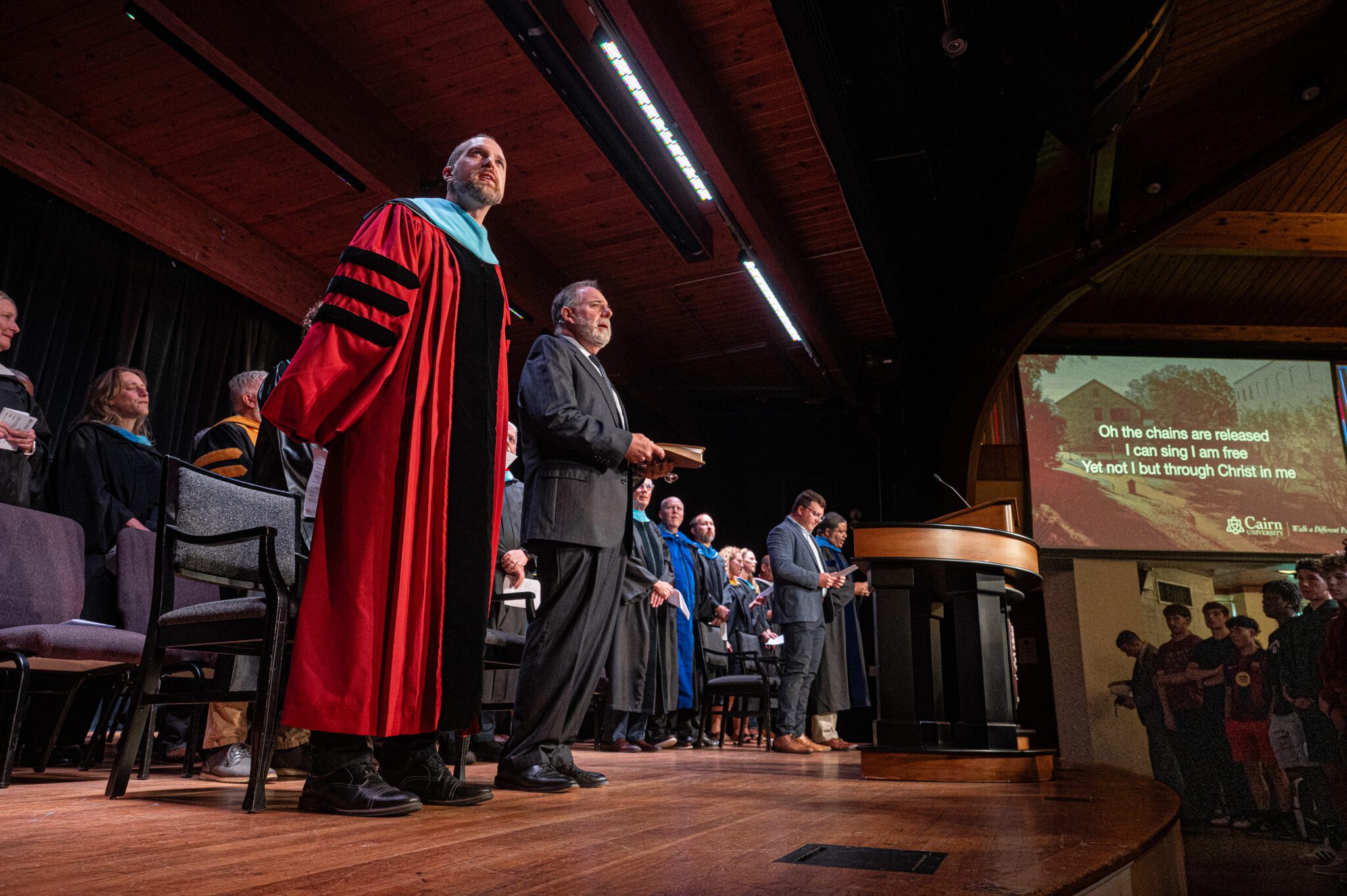 University President and Provost standing on chapel stage singing with faculty behind them